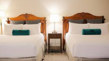 Habitación de hotel con dos camas, cada una con cabecera de madera, ropa de cama blanca y almohadas estampadas. Una mesita de noche con lámpara se encuentra entre las camas.