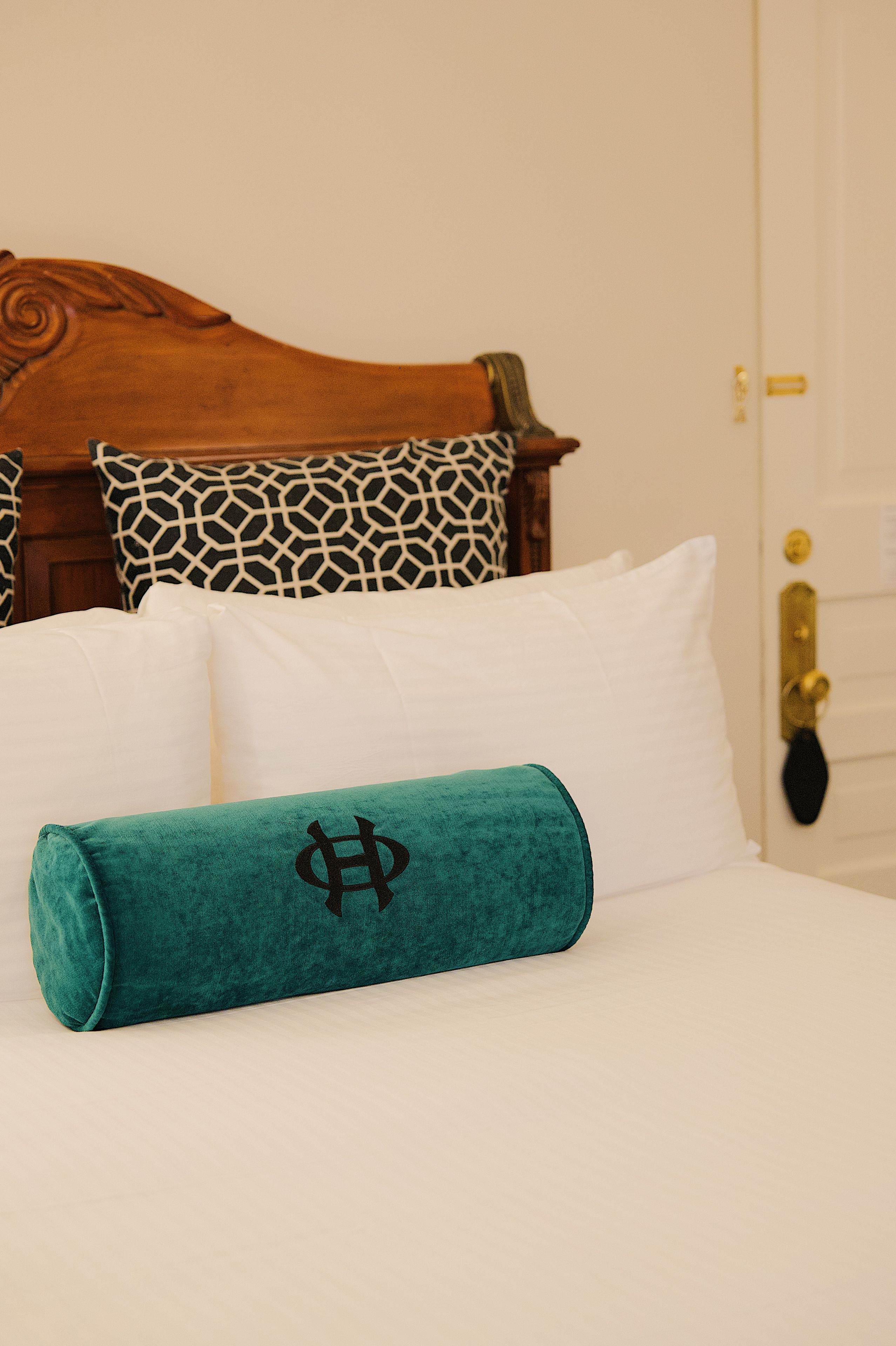 A bed with white bedding, a teal pillow with a symbol, and a patterned pillow.