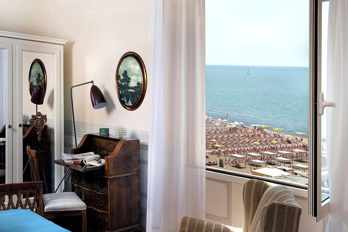 A hotel room with a view of a beach, a desk with a lamp, a chair, and a bed.