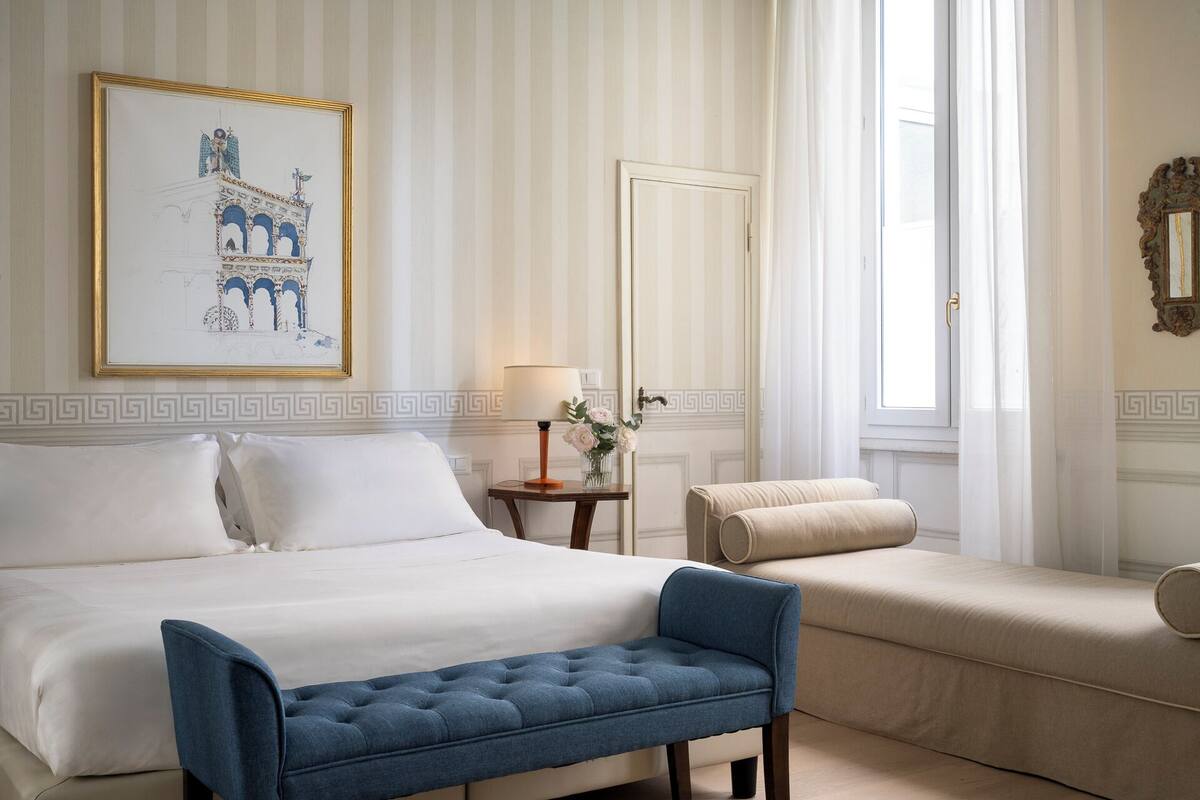 A hotel room with a large bed, a blue bench, a bedside table with a lamp and flowers, a door, and a framed picture on the wall.
