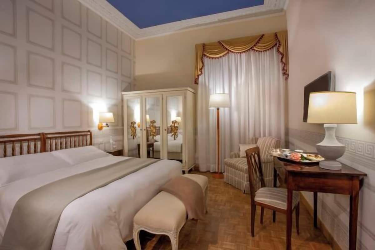 A hotel room with a large bed, a desk with a lamp, a chair, a wardrobe, and a mirror.