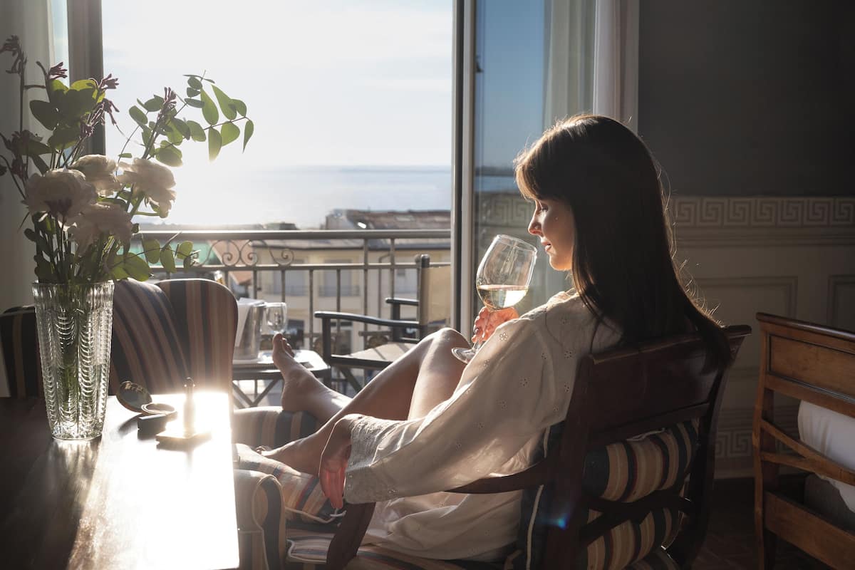 A woman sitting by a window with a view of the sea, holding a glass of wine, with a vase of flowers on the table.