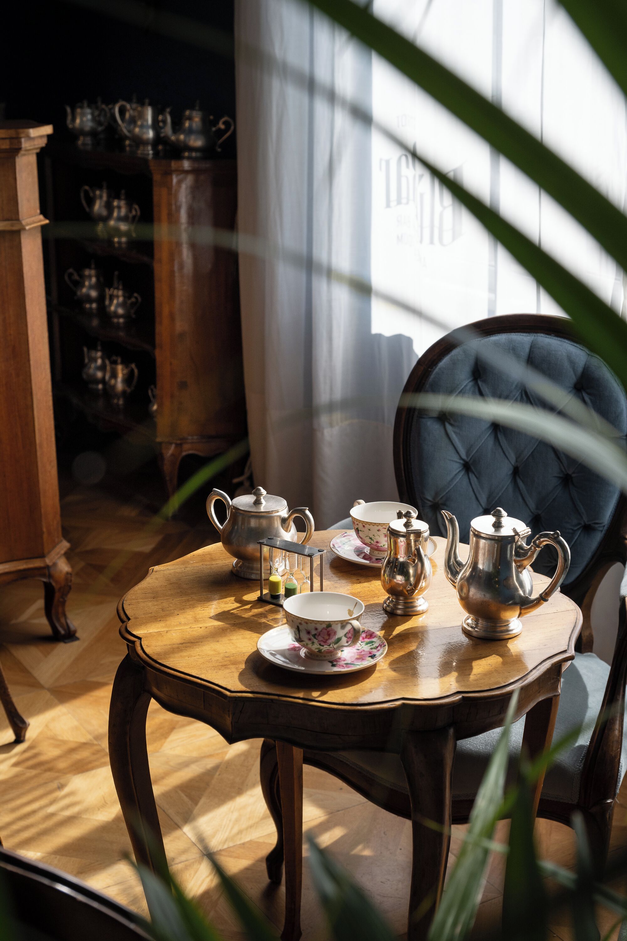 Una mesa de madera con juego de té y una silla tapizada en azul en una habitación con suelo de madera y un armario al fondo.