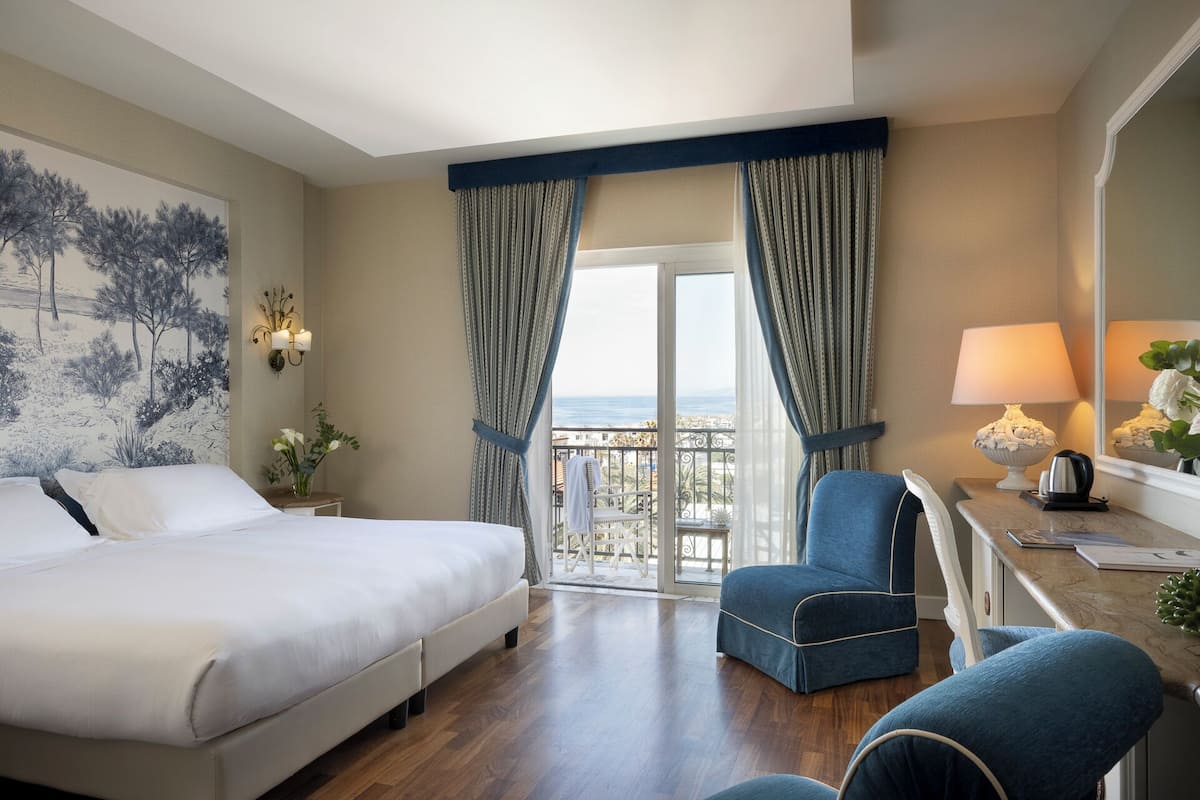 A hotel room with a bed, a desk with a lamp, a chair, and a view of the sea through large windows.