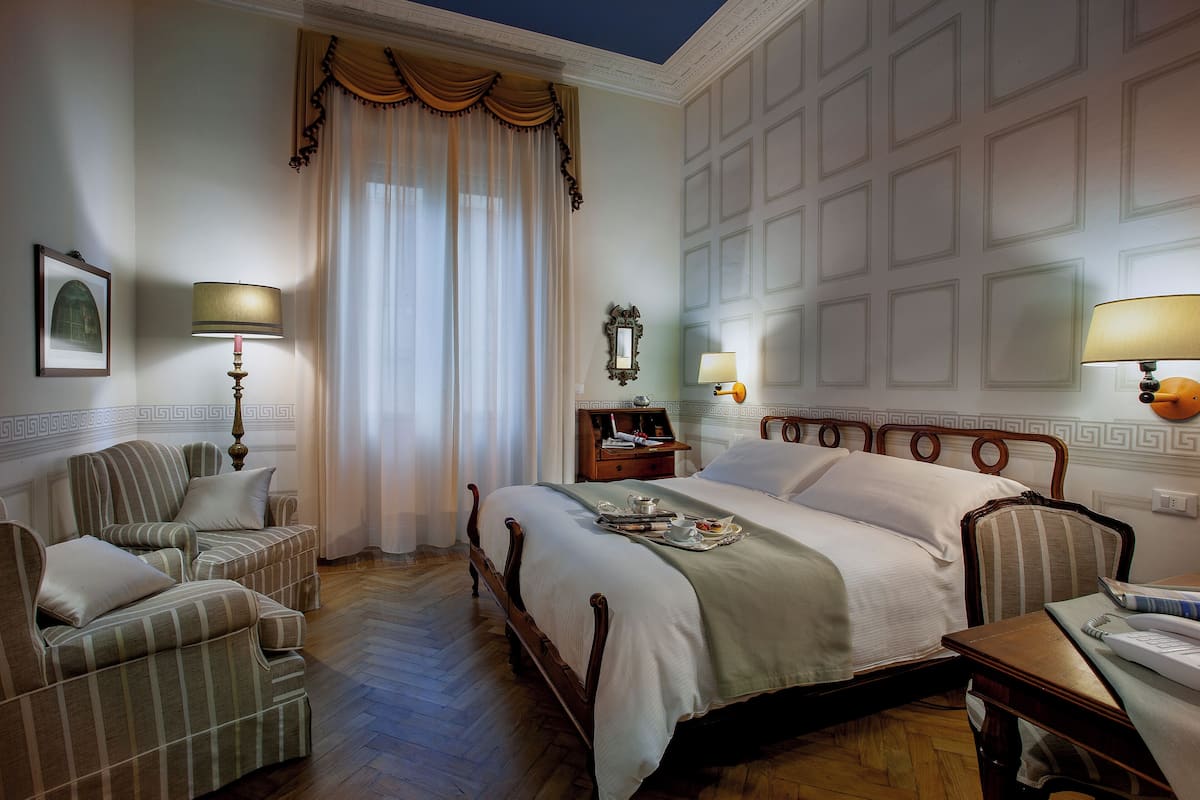 A hotel room with a large bed, two armchairs, a wooden nightstand, and a table with a lamp.