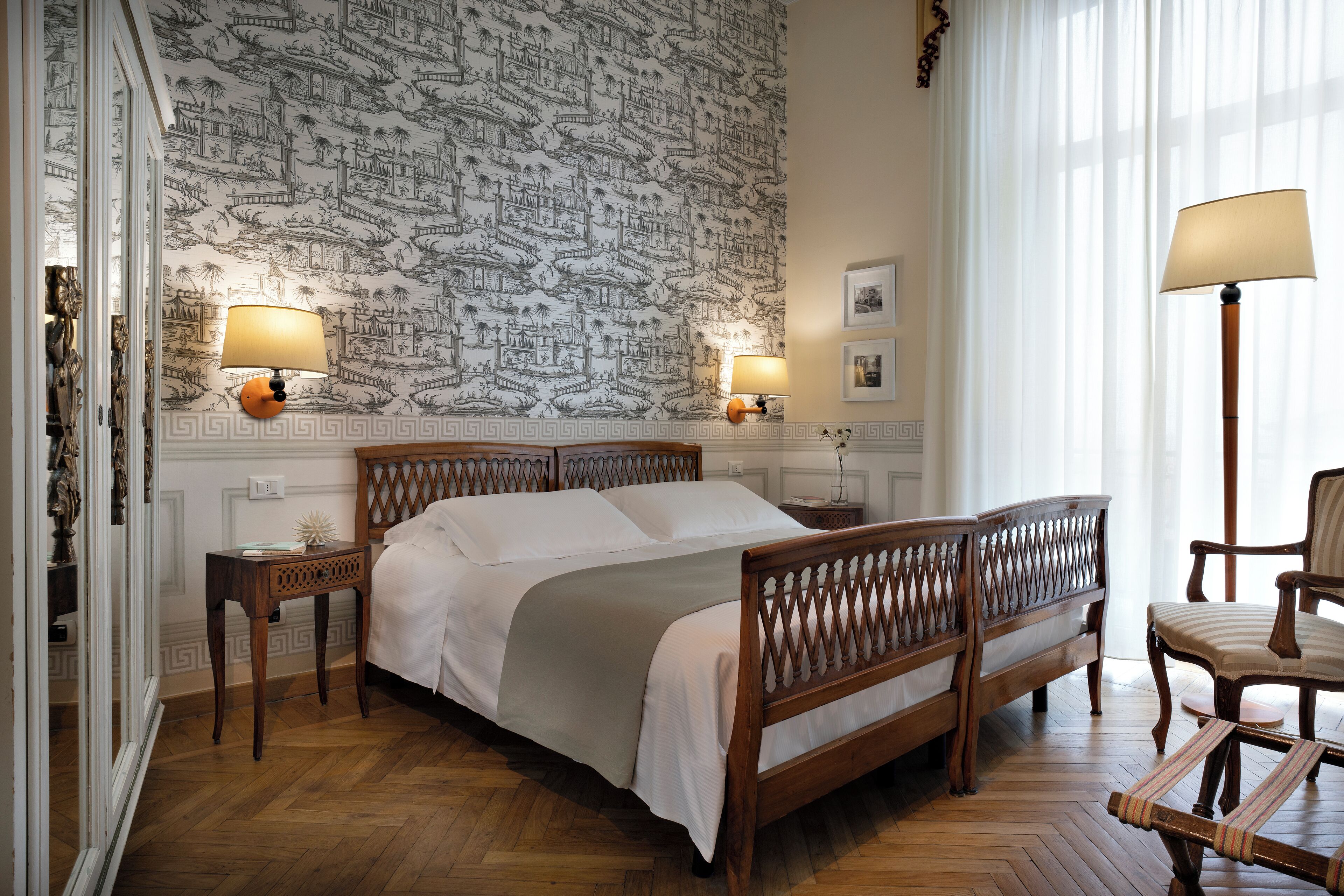 A bedroom with a wooden bed, two bedside tables with lamps, a chair, and a patterned wallpaper.