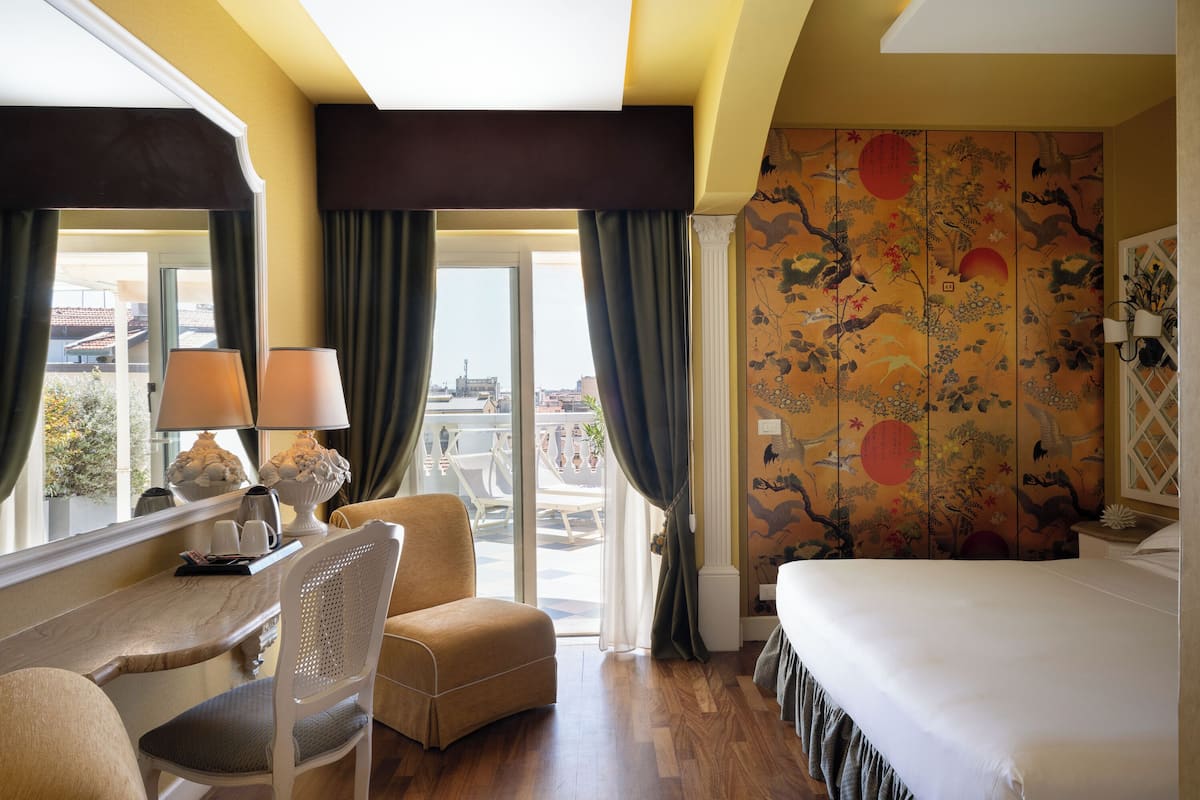 A hotel room with a bed, a desk with a chair, a window with curtains, and a view of the city.