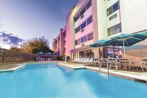 A swimming pool with lounge chairs and umbrellas, surrounded by a building with multiple floors and windows.