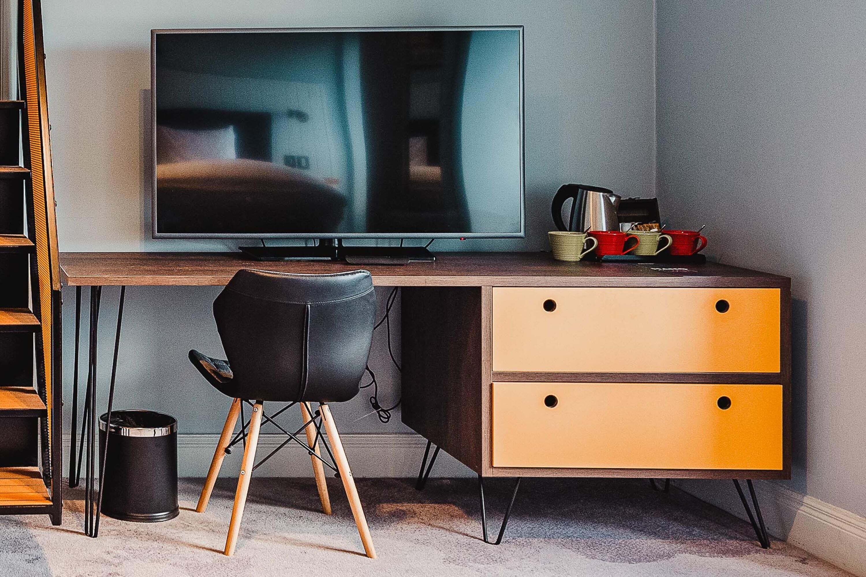 Ett modernt rum med en platt-TV på ett skrivbord i trä, en svart stol, en gul byrå och en svart papperskorg.