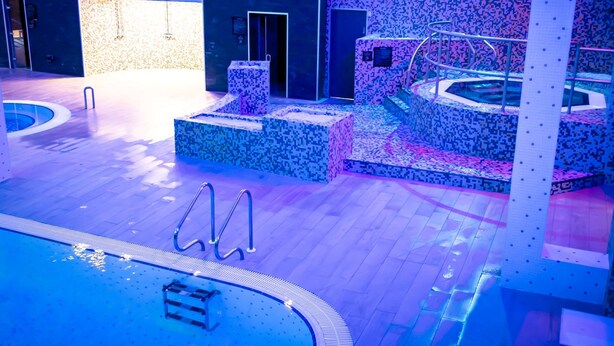 Un área de piscina interior moderna con un jacuzzi central, paredes con azulejos y suelo de madera.