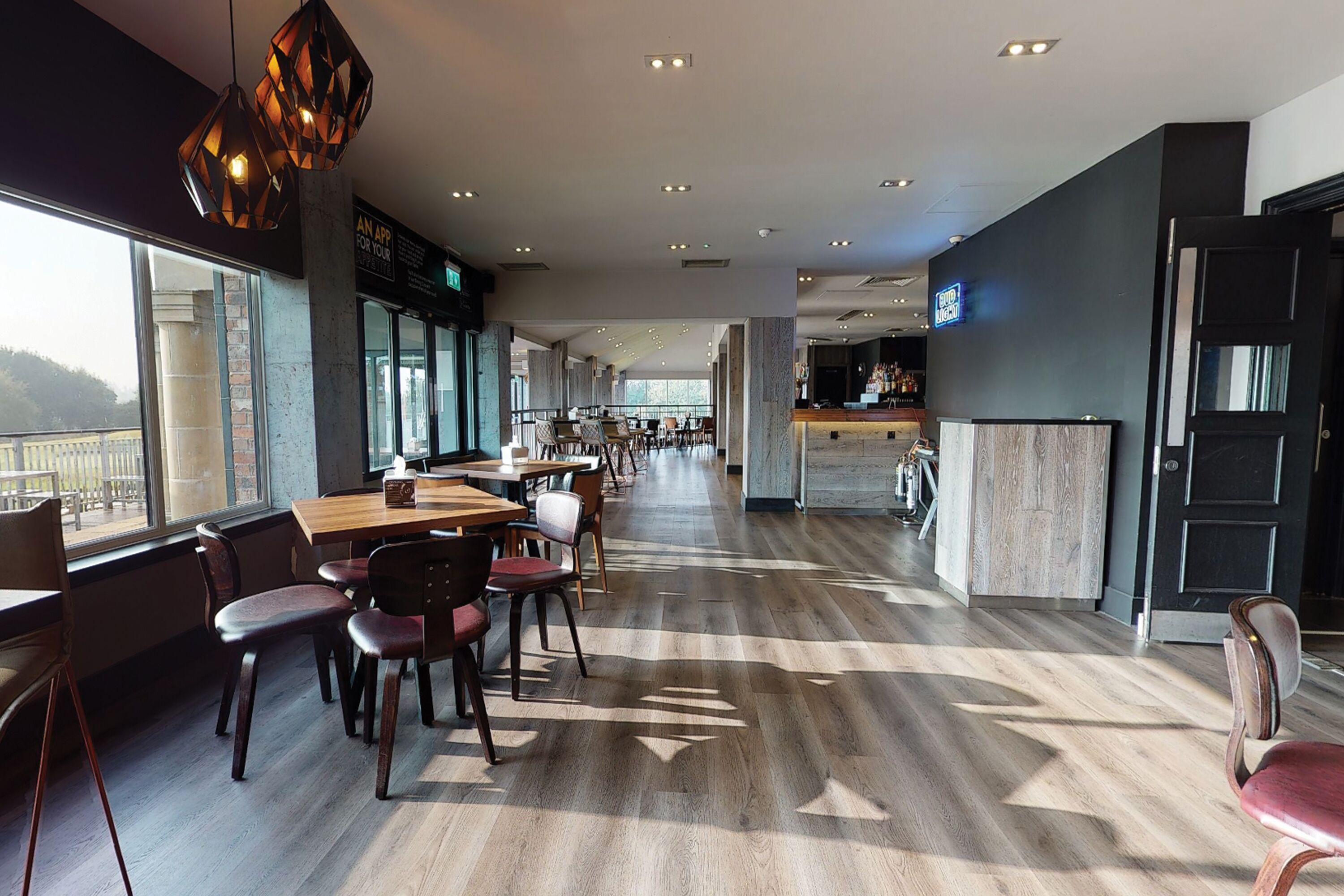 A modern restaurant with wooden flooring, tables, and chairs. There is a bar area in the background.