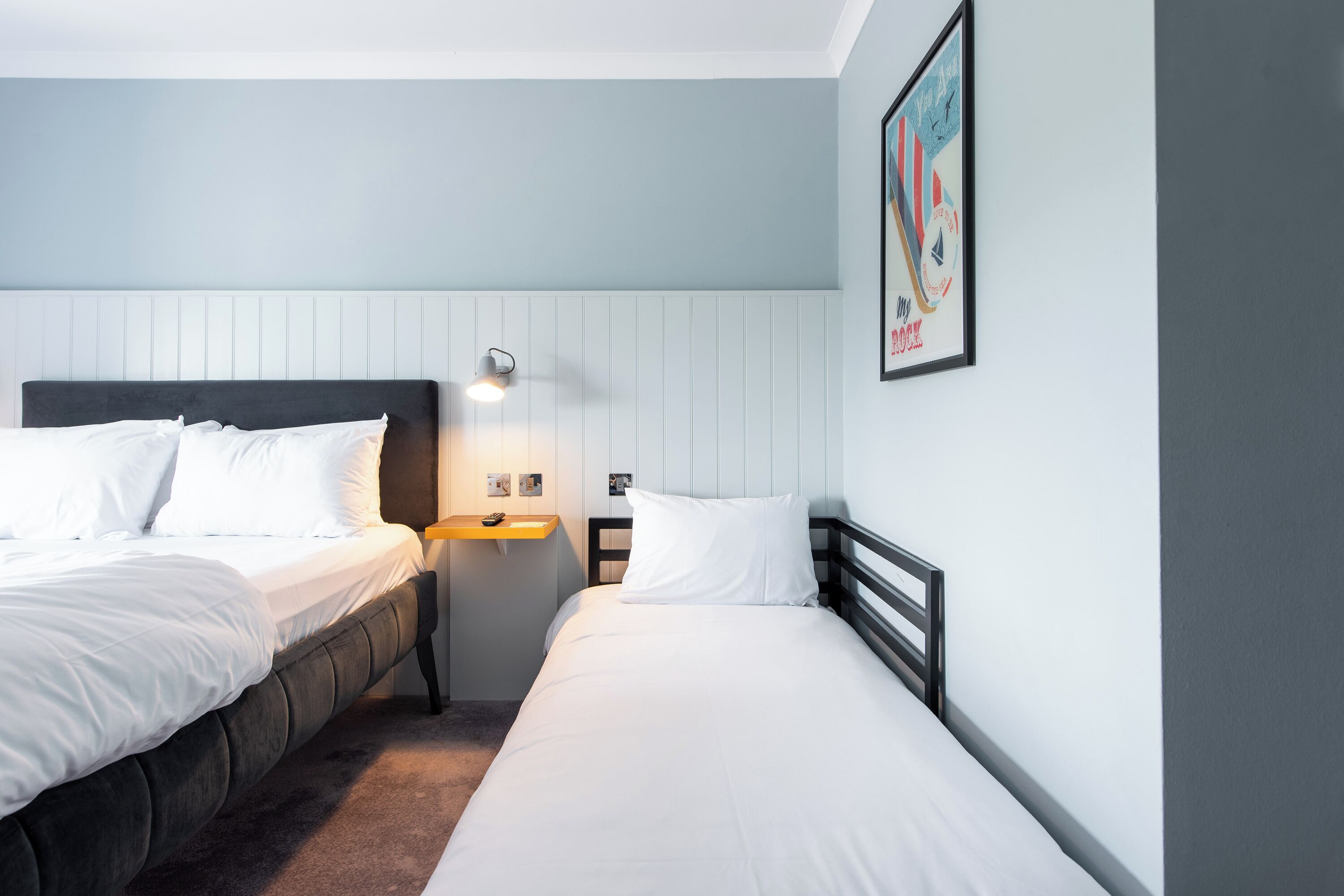 A hotel room with two beds, a bedside table, a wall-mounted light, and a framed picture.