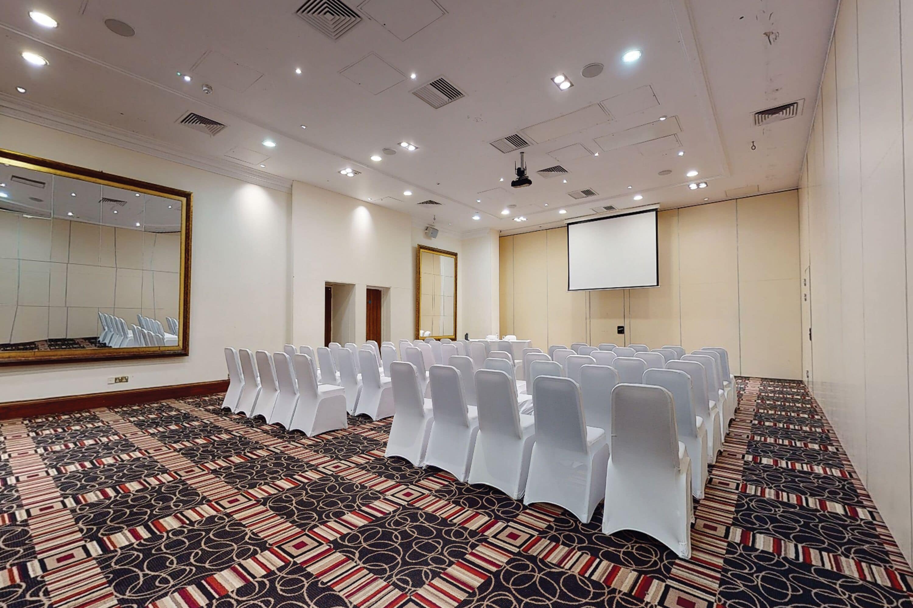 A conference room with rows of white chairs, a large projection screen, and a patterned carpet.