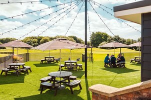Een terras buiten met picknicktafels en parasols, omgeven door een hek en groen.