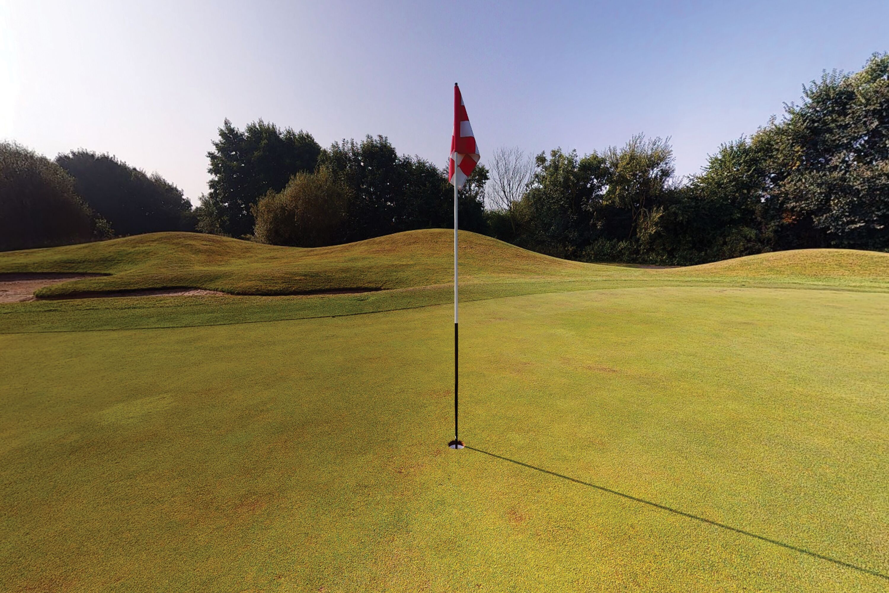 A golf course with a flag on a flagpole, surrounded by rolling hills and trees.