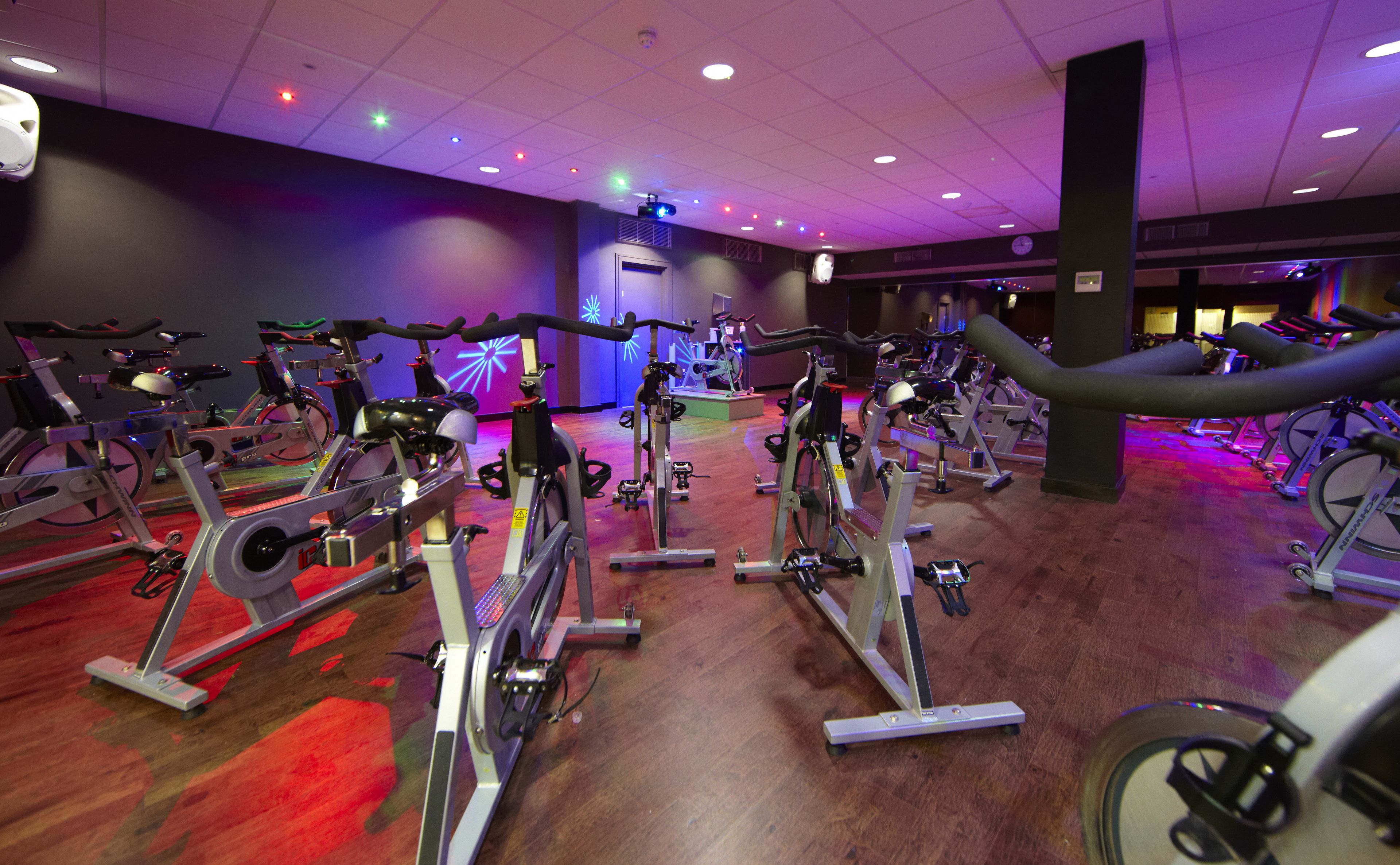 A gym with multiple stationary bikes and elliptical machines, a wooden floor, and a ceiling with recessed lighting.