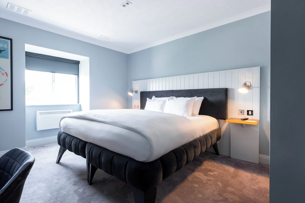 A modern bedroom with a large bed, a bedside table, a chair, and a window with blinds.