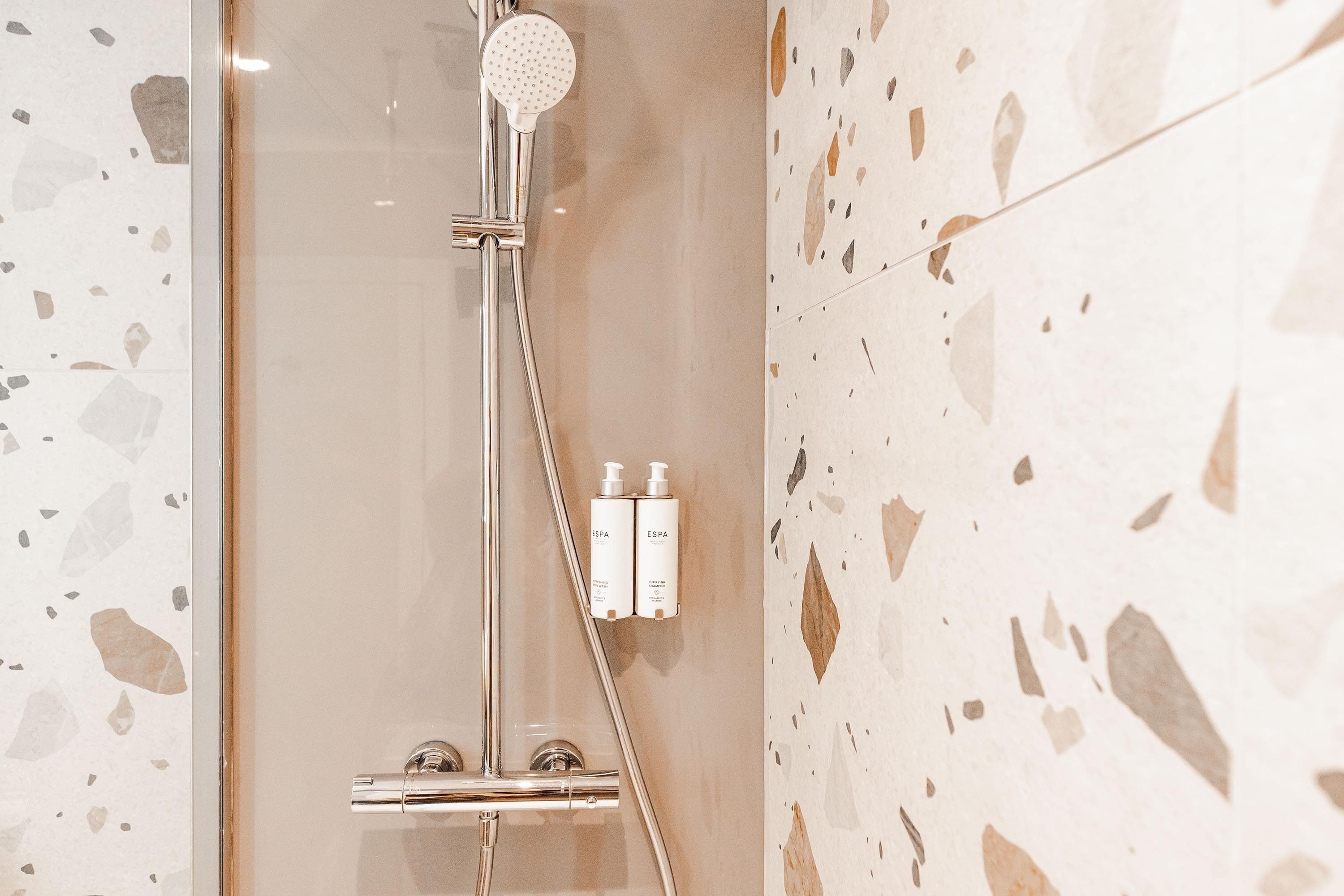 Un baño moderno con ducha acristalada, cabezal de ducha manual y dos botellas de champú en la pared.