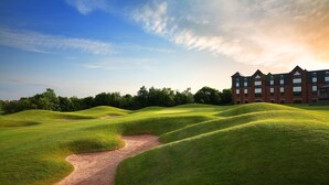 Un campo de golf con colinas verdes ondulantes, un búnker de arena y un edificio de ladrillo grande al fondo.