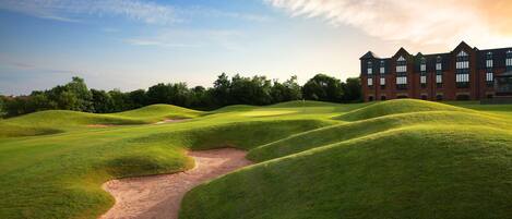 Un parcours de golf avec des collines verdoyantes, un bunker de sable et, au loin, un grand bâtiment en briques.