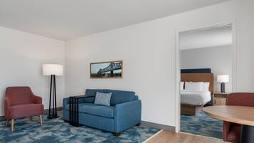 Habitación de hotel con sofá azul, sillón rojo, una mesita, lámpara, cama con cabecero blanco y un cuadro de un puente.