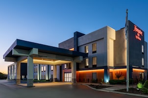A modern hotel with a clear entrance, a sign indicating it's a Hampton Inn by Hilton, and a covered area with a 'Welcome' sign.