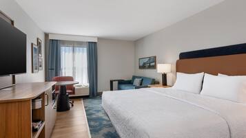 A hotel room with a bed, a desk, a chair, a television, a window with curtains, a painting on the wall, and a lamp.