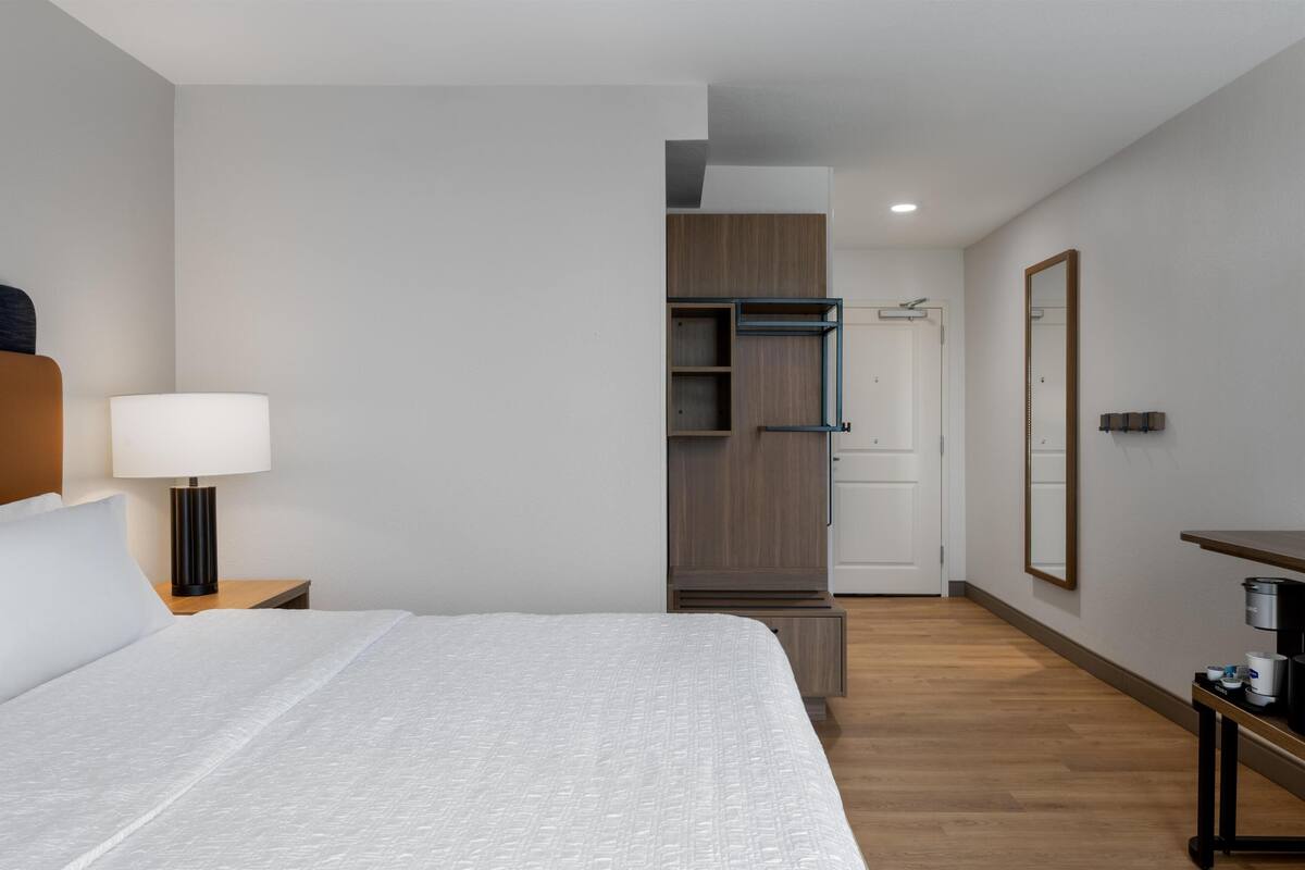 A hotel room with a bed, a nightstand with a lamp, a wardrobe, a mirror, and a small table with a coffee maker.