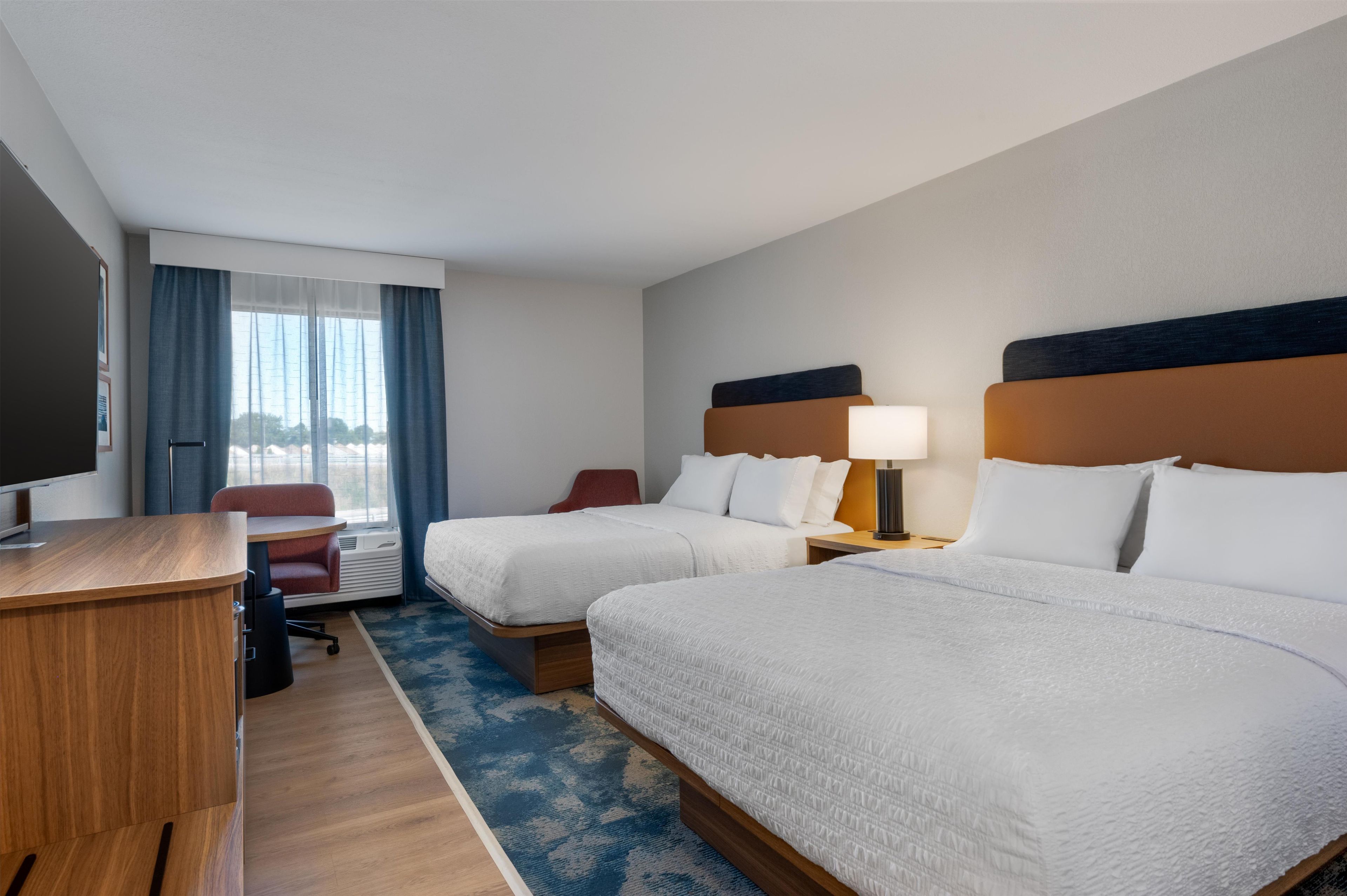 Habitación de hotel con dos camas, un escritorio, una silla, un televisor y una ventana con cortinas.