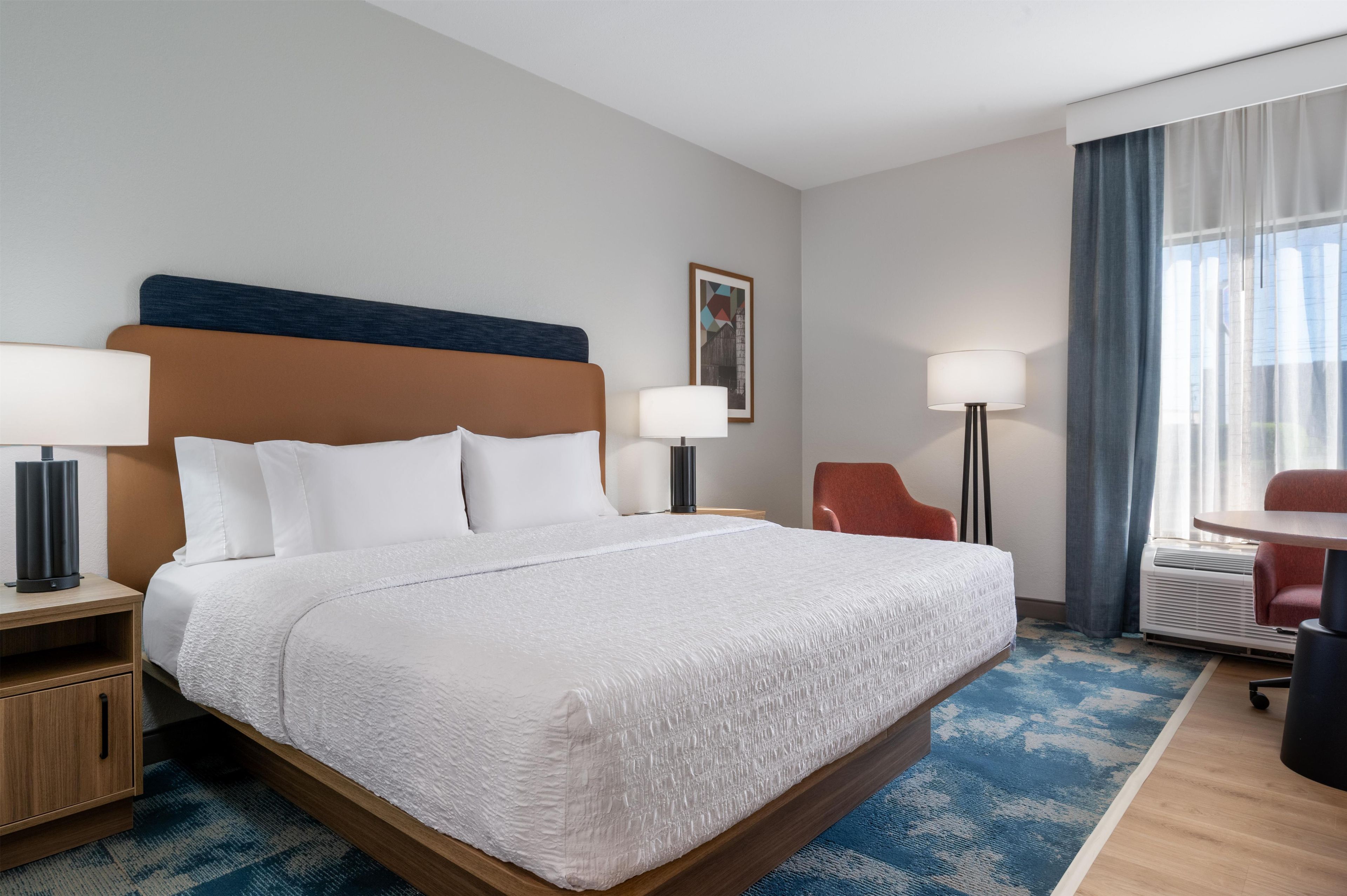 A hotel room with a large bed, two bedside tables, a desk, a chair, a lamp, and a window with curtains.