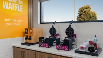 A waffle station with two waffle makers, a sign for malted vanilla and s'mores waffle batter, and a container for strawberry topping.