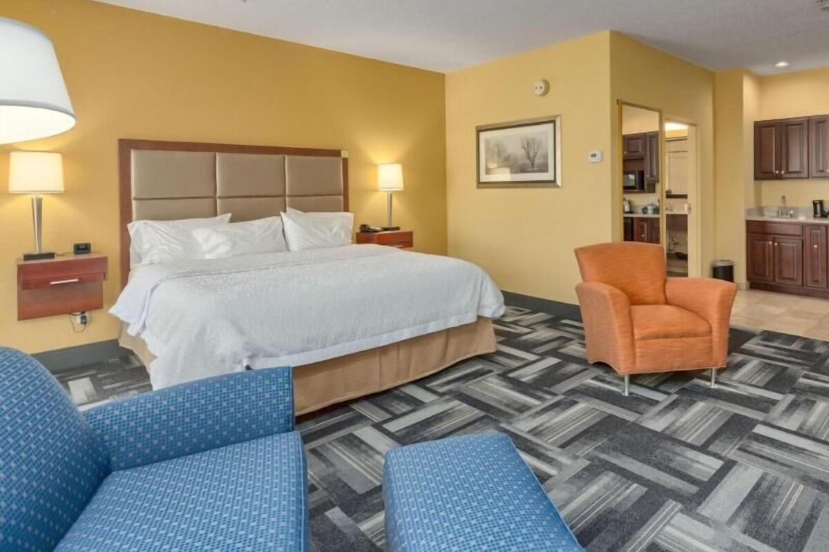 Ein Hotelzimmer mit einem Bett, zwei Nachttischen mit Lampen, einem Sessel, einem kleinen Tisch und einer Küchenzeile im Hintergrund.