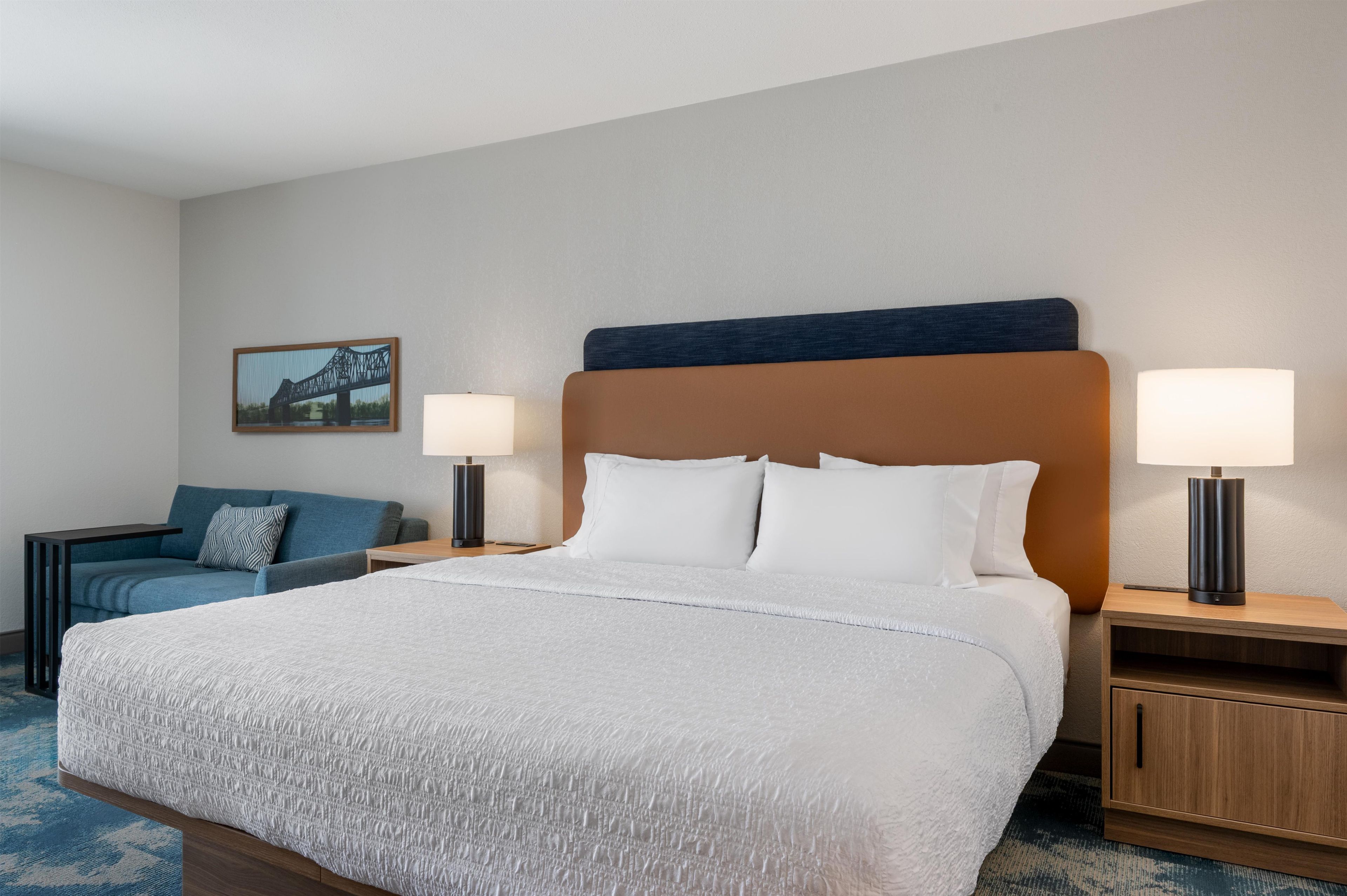 A hotel room with a large bed, two bedside tables with lamps, a blue sofa, and a framed picture on the wall.
