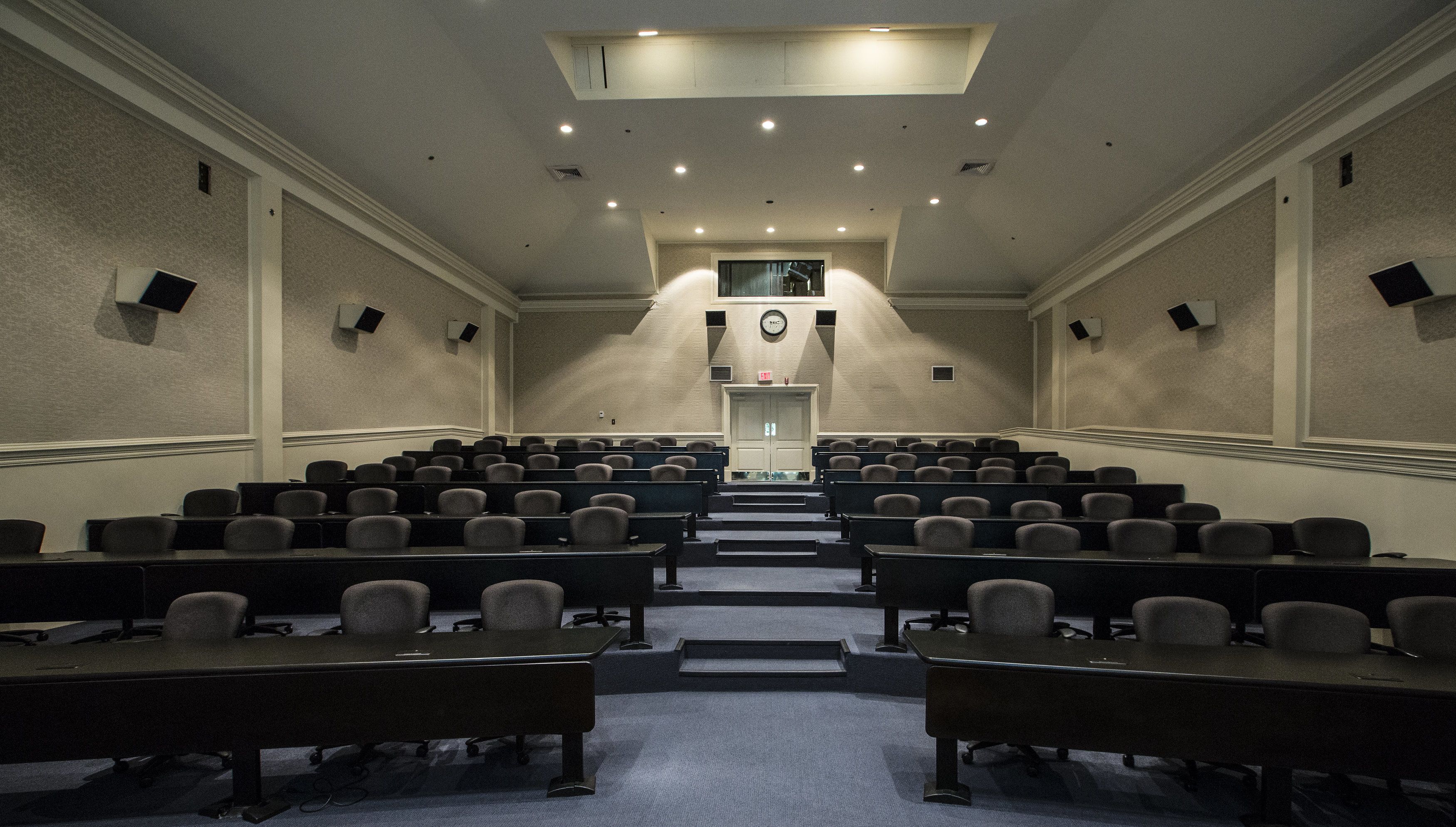 A conference room with rows of chairs and tables, a clock on the wall, and a staircase leading to an upper level.