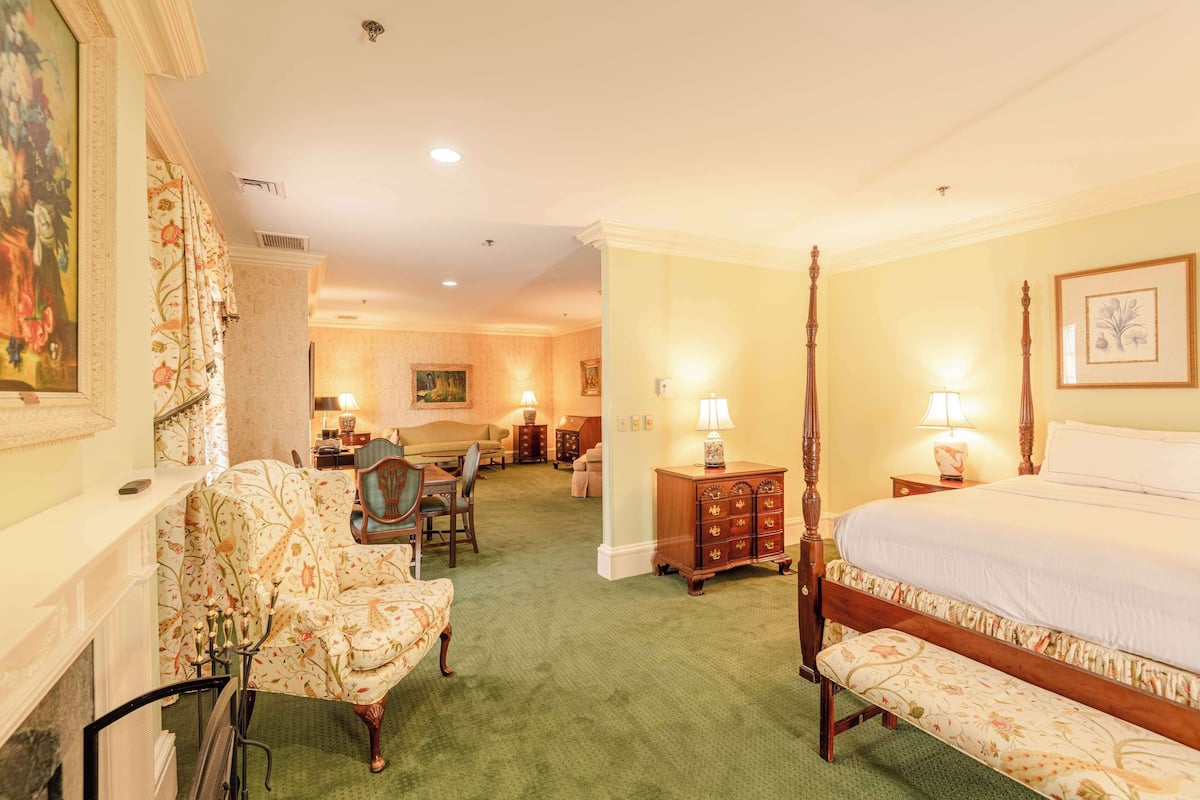 A hotel room with a four-poster bed, a sitting area with a sofa and chairs, a wooden cabinet, and a dining table with chairs.