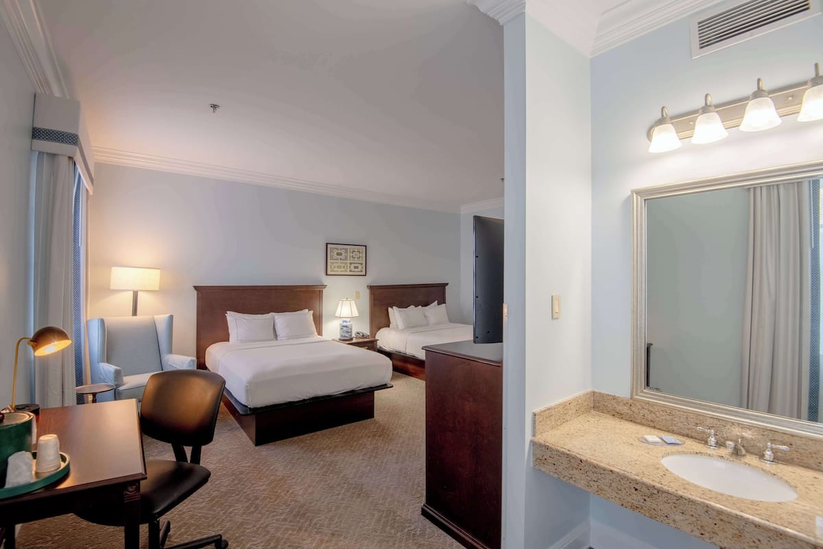 A hotel room with two beds, a desk with a chair, a vanity with a mirror, and a bathroom with a sink and mirror.