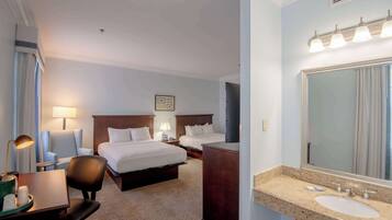 A hotel room with two beds, a desk with a chair, a vanity with a mirror, and a bathroom with a sink and mirror.