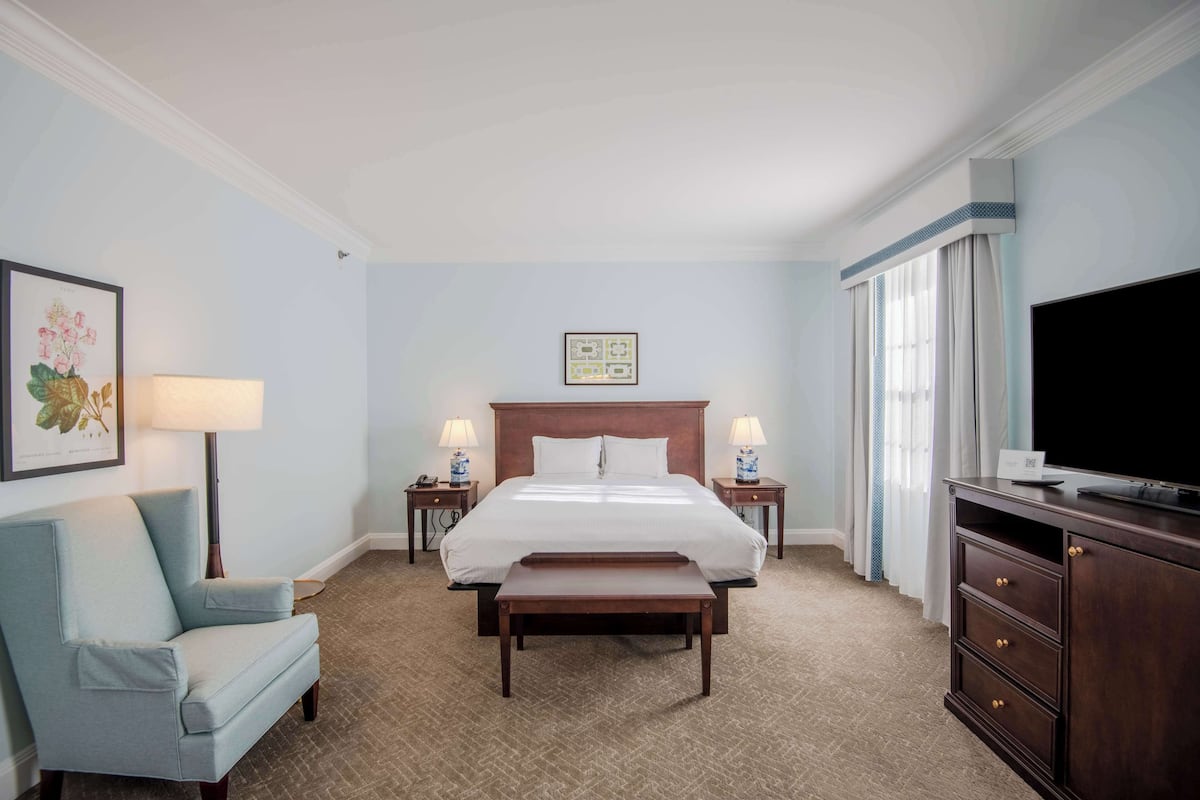 A hotel room with a large bed, a bedside table with a lamp, a chair, a bench, a dresser, and a TV.