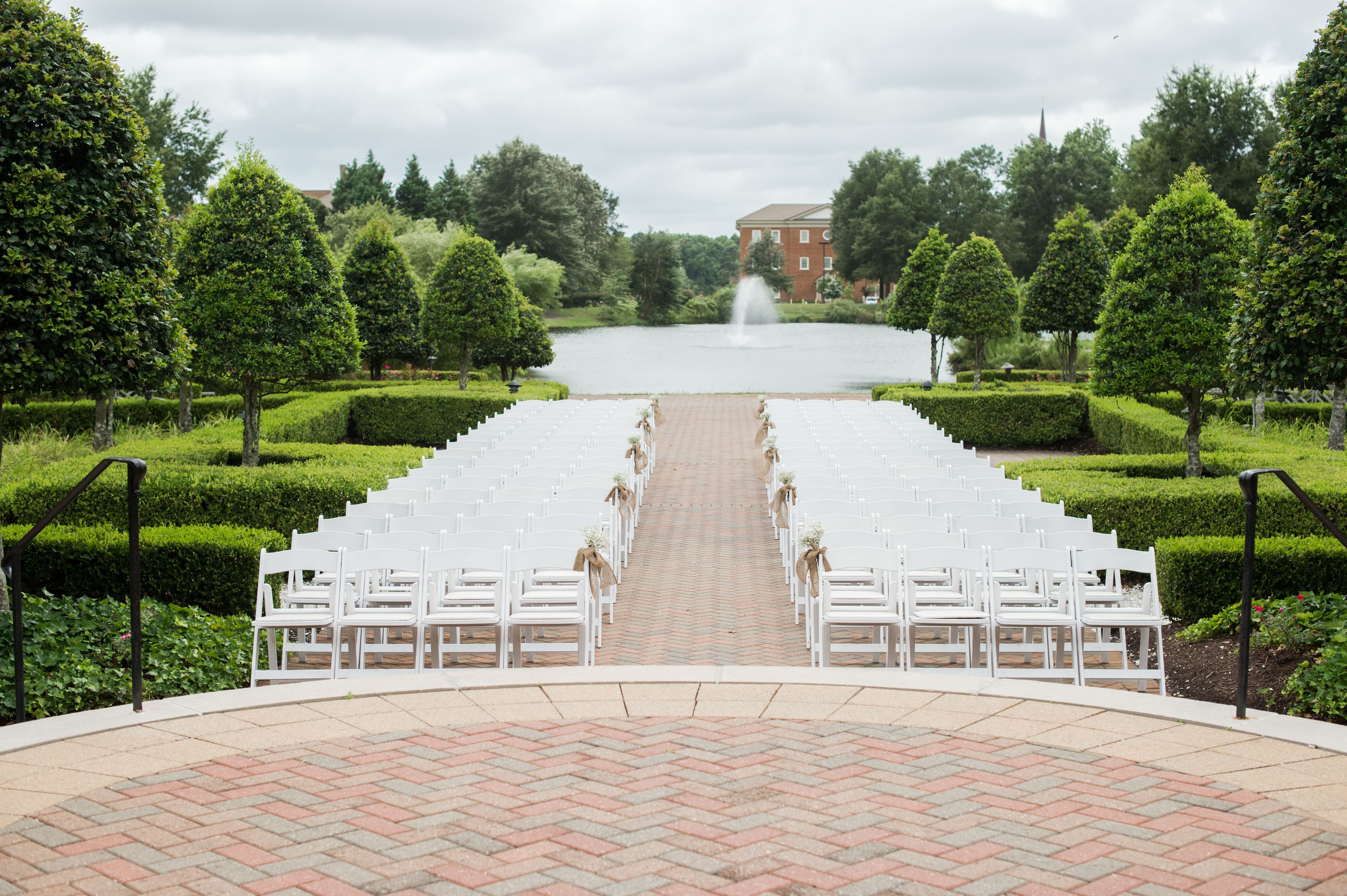 Een formeel buitengebied met rijen witte stoelen, klaargezet voor een evenement, een met bakstenen bestraat pad dat naar een waterpartij leidt, en een gebouw op de achtergrond.