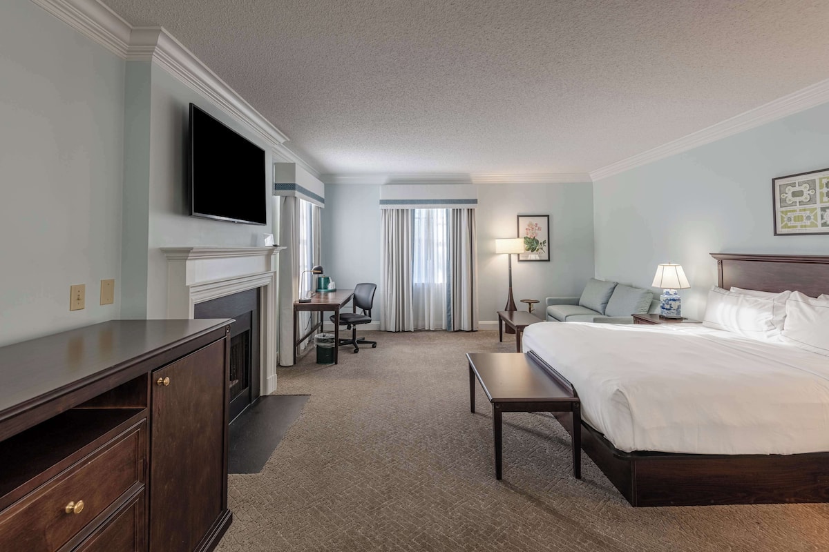 A hotel room with a large bed, a desk with a chair, a TV mounted on the wall, and a fireplace.