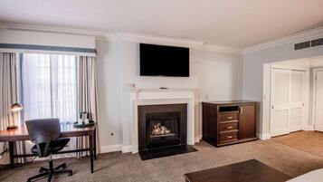 A hotel room with a fireplace, a flat-screen TV mounted above it, a desk with a chair, and a large bed.