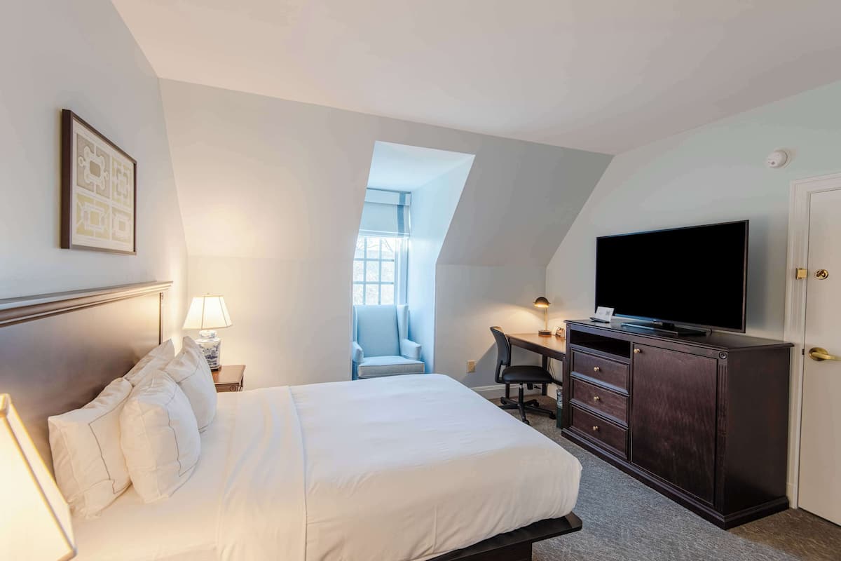 A bedroom with a bed, a desk, a chair, a TV, and a window.