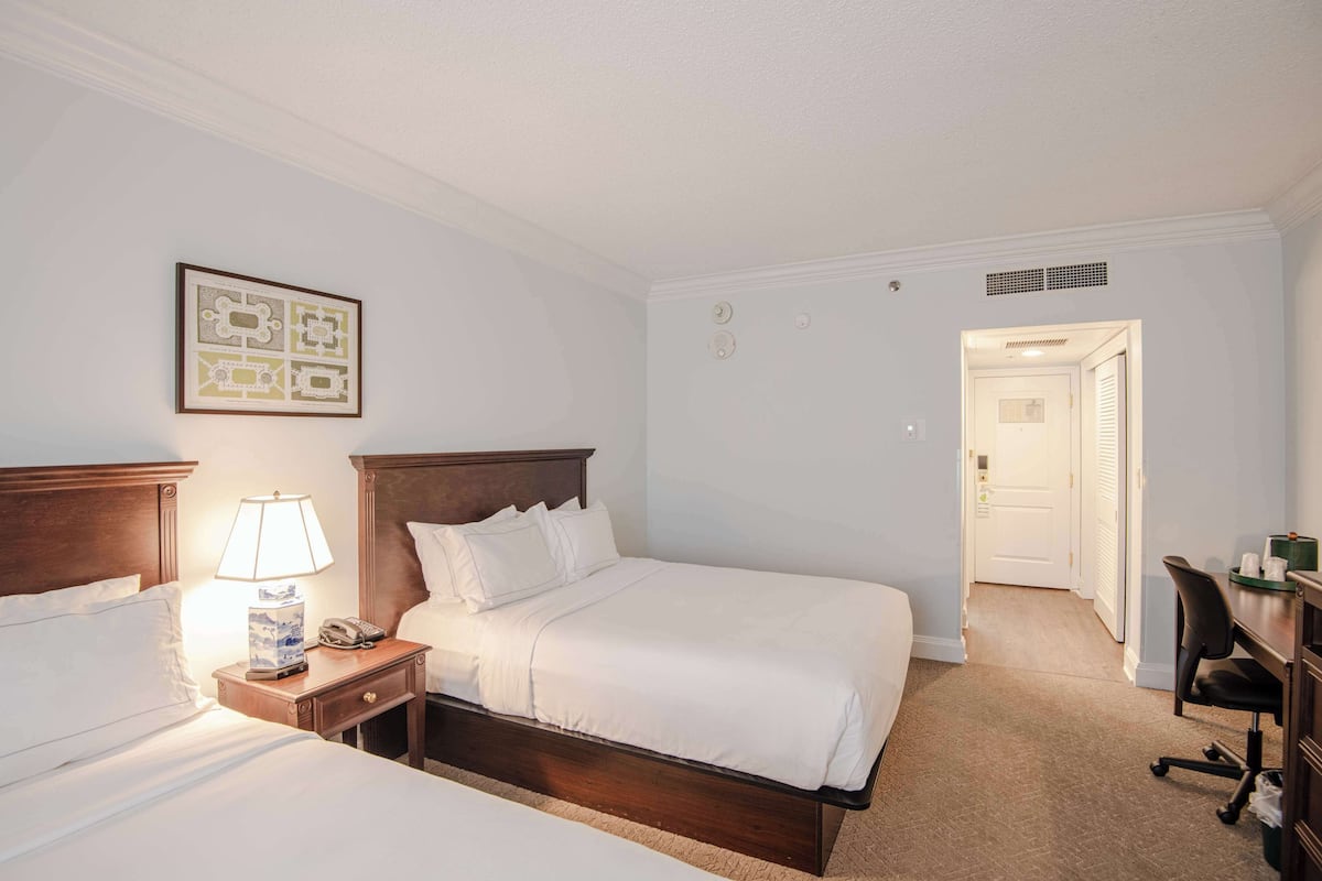 A hotel room with two beds, a desk, a chair, a lamp, and a framed picture on the wall.