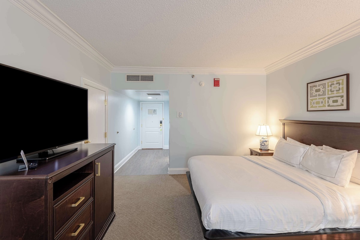 A hotel room with a large flat-screen TV on a wooden dresser, a neatly made bed with white linens, a bedside lamp, and a framed picture on the wall.
