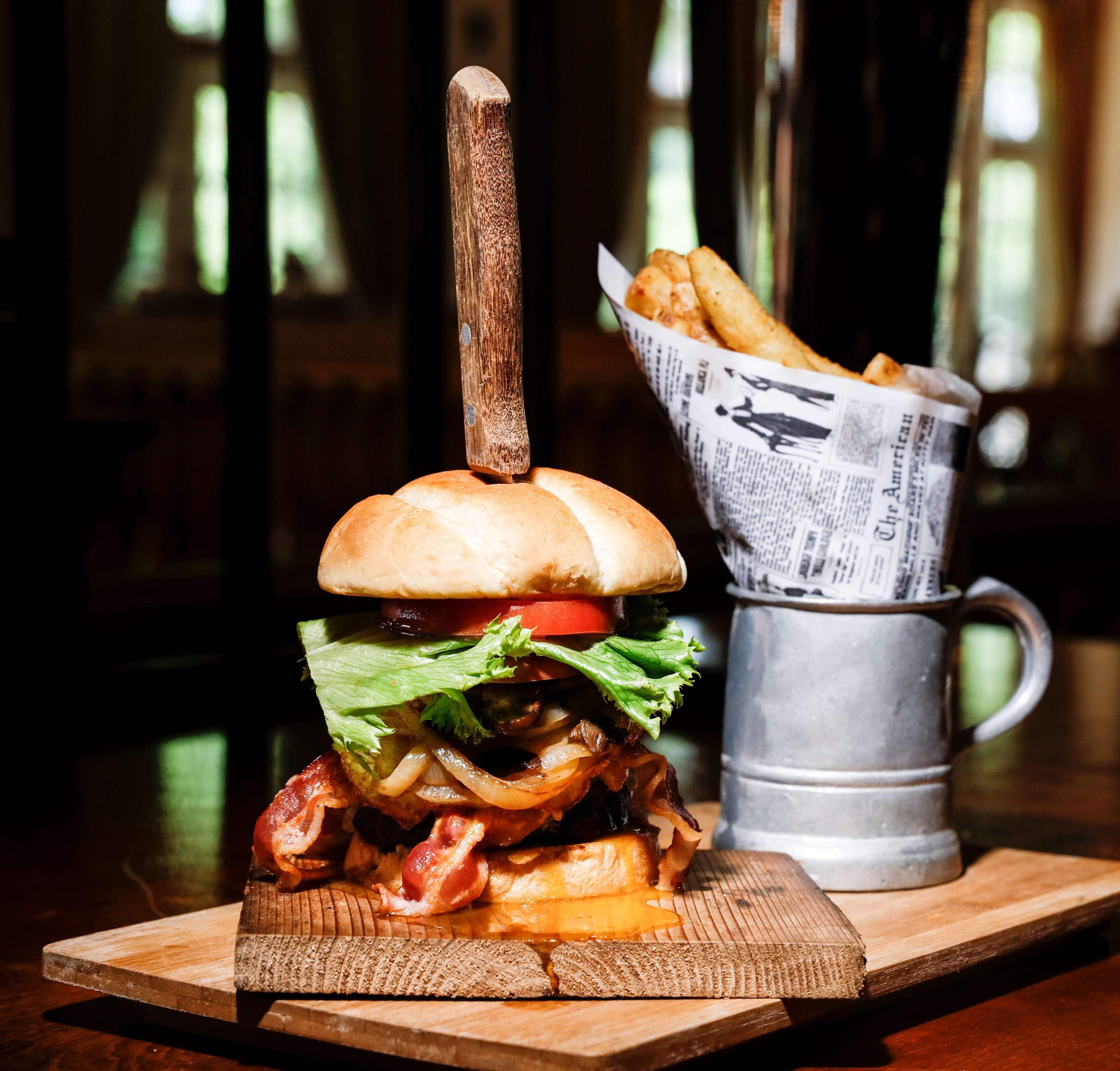 Una hamburguesa con un palillo de madera sobre una tabla de madera, un vaso metálico y una cesta de papas fritas.