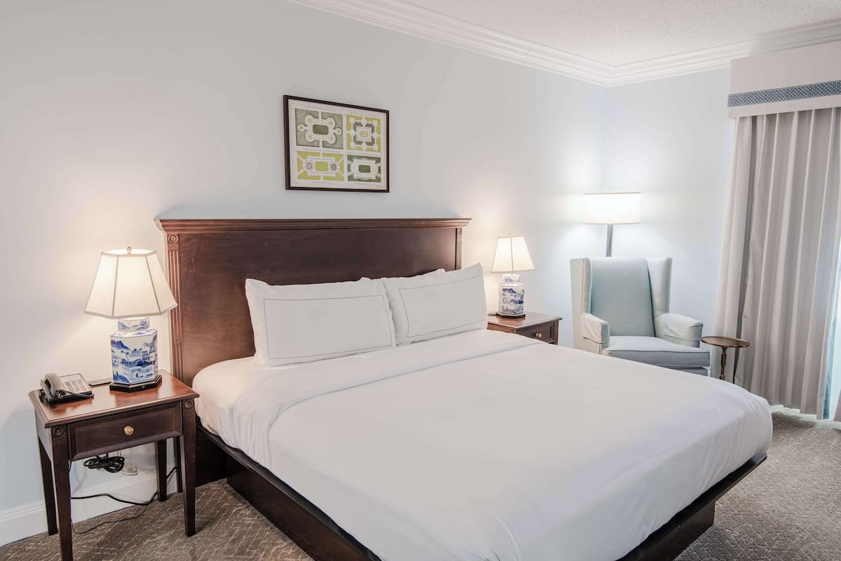 A neatly made bed with white linens, two bedside tables with lamps, a chair, a framed artwork on the wall, and a window with curtains.
