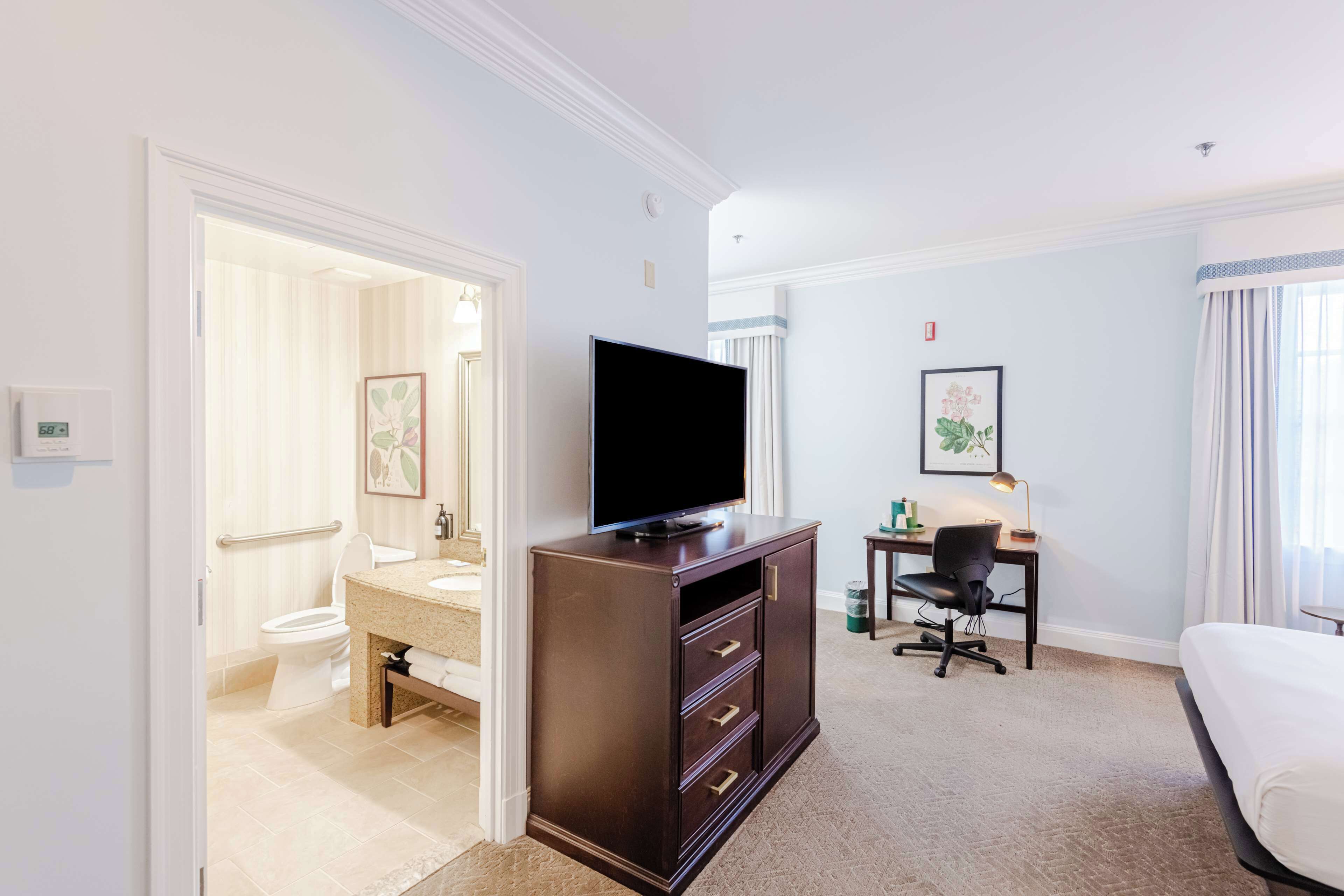 A hotel room with a bed, a desk with a chair, a television on a dresser, a bathroom with a toilet and sink, and a framed picture on the wall.