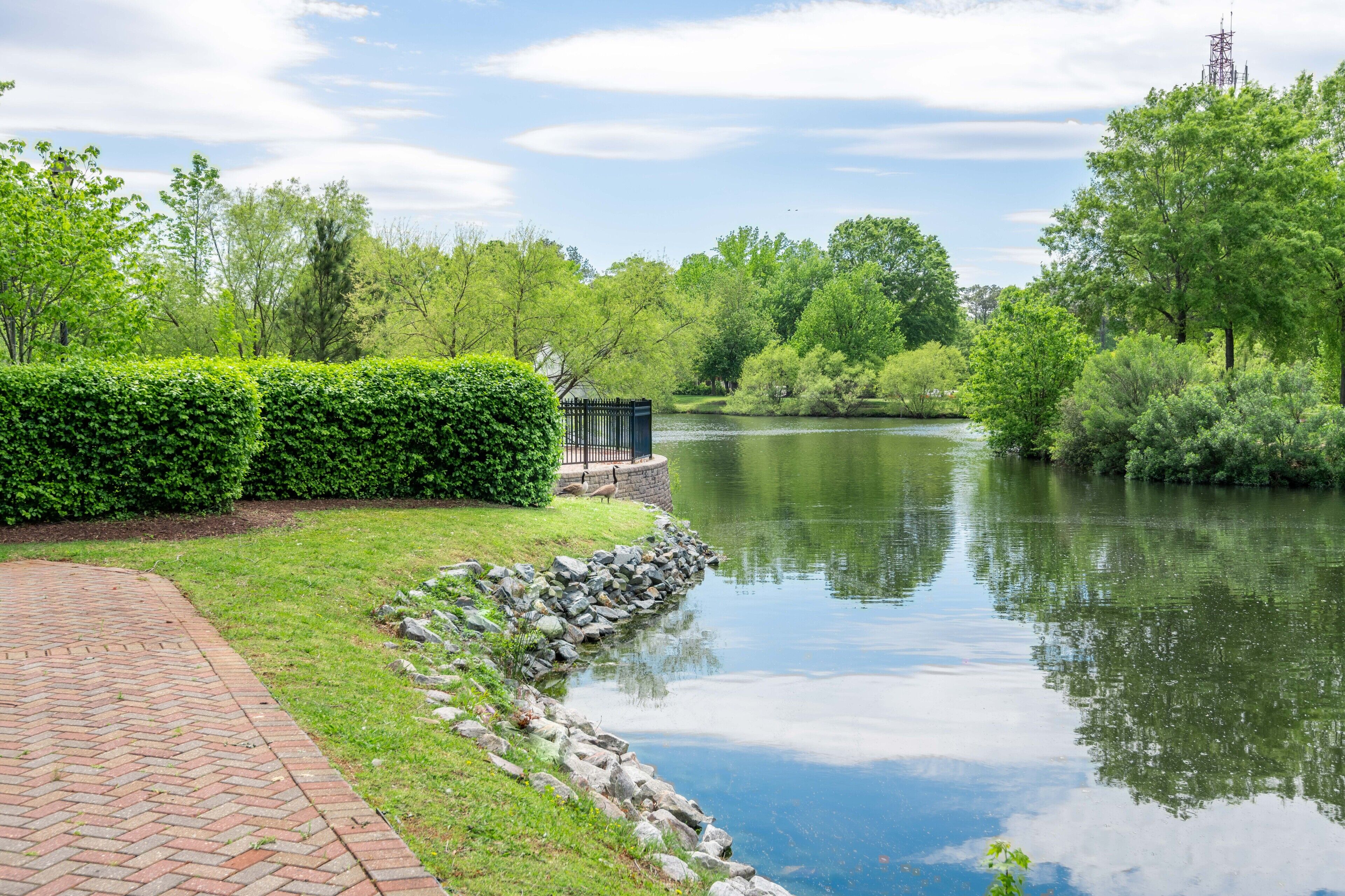 A serene riverside pathway with a brick walkway, lush greenery, and a small structure near the water.