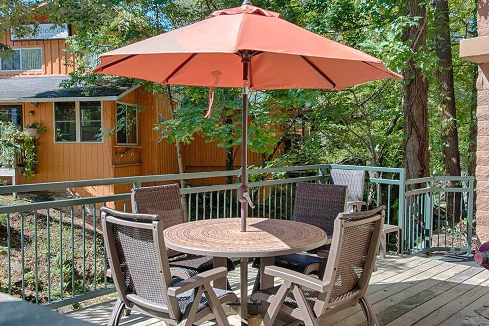 A round table with wicker chairs and a red umbrella on a wooden deck.