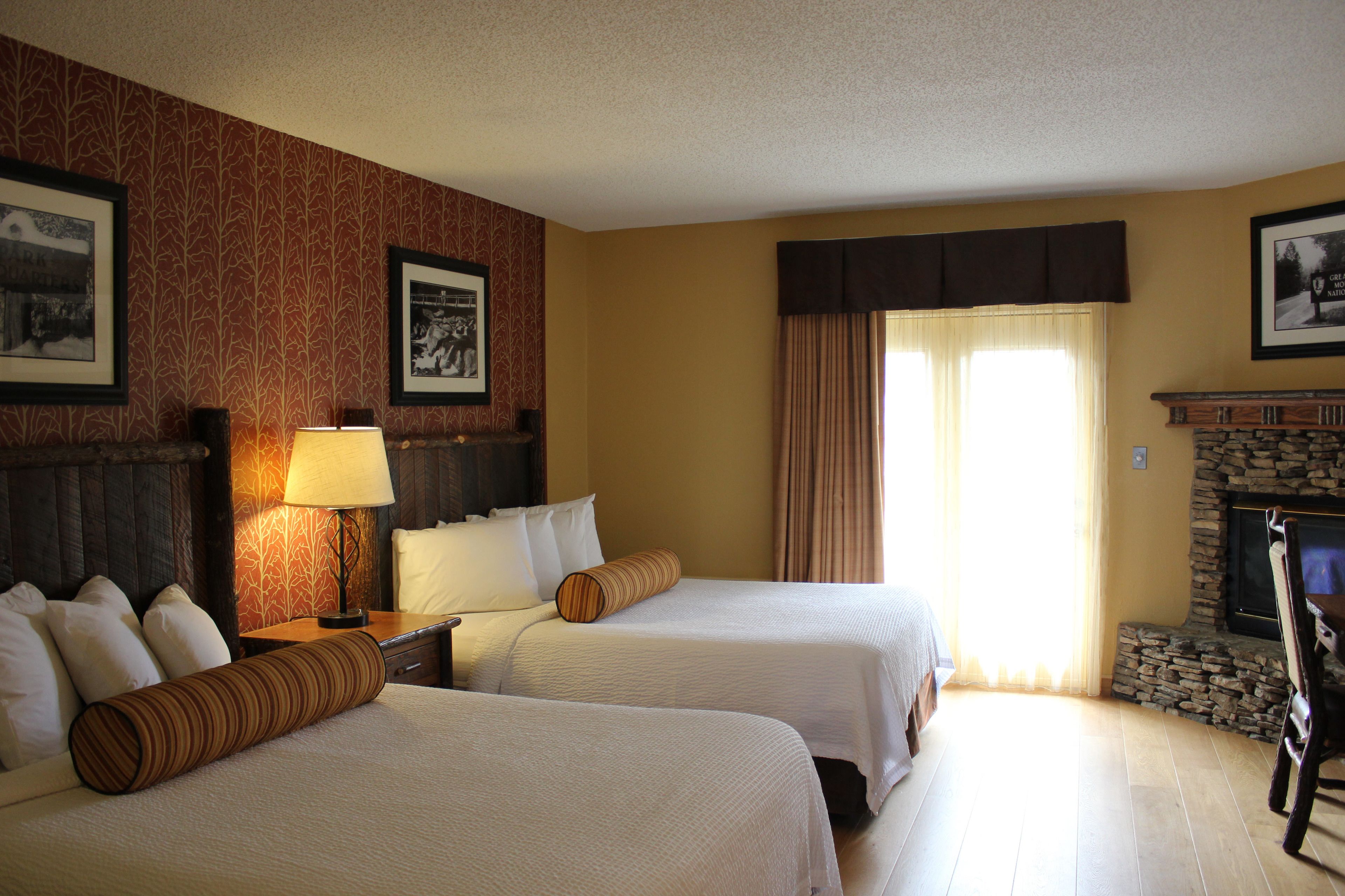 A hotel room with two beds, a nightstand, a lamp, a fireplace, and framed pictures on the walls.